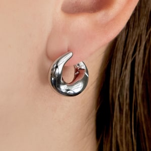 May include: A close-up of a silver-colored hoop earring. The earring has a unique, curved design with a smooth, reflective surface. The earring is worn on an ear, with the person's hair and skin visible in the background.