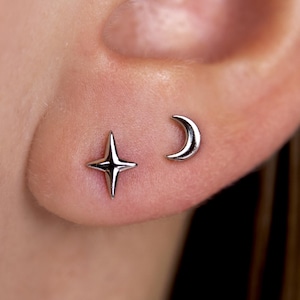 May include: A close-up of an ear with two small, silver earrings. One earring is a four-pointed star, and the other is a crescent moon. The earrings are simple and elegant, likely made of sterling silver.