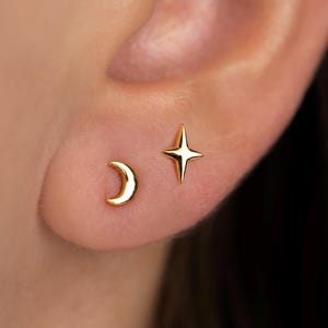 May include: A close-up of a person's ear with two small, gold-colored stud earrings. One earring is a crescent moon shape, and the other is a four-pointed star. The earrings are shiny and reflect light.
