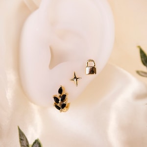 May include: Three gold earrings with black accents. The first earring is a small star, the second is a padlock, and the third is a cluster of black stones.