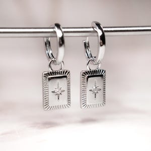 May include: A pair of silver hoop earrings with rectangular pendants. Each pendant features a starburst design with a small, clear gemstone in the center. The earrings are displayed on a silver bar against a soft, blurred background.