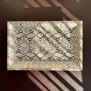 Könnte beinhalten: Ein rechteckiges Tablett mit einem Schlangenhautmuster in Beige- und Schwarztönen. Das Tablett ist aus einem harten Material gefertigt und hat eine glatte Oberfläche.