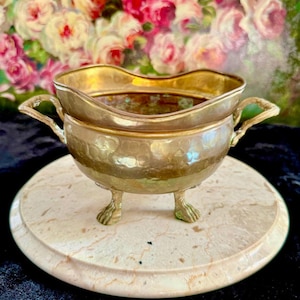 Puede incluir: Un pequeño cuenco antiguo de color plateado con dos asas y tres patas de garra. El cuenco se asienta sobre una base redonda, similar al mármol. El interior del cuenco es de un color más oscuro y el borde es dorado. El fondo presenta una pintura floral.