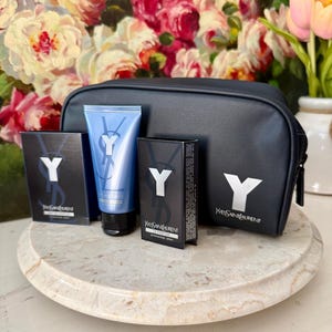 May include: A black cosmetic bag and matching products from Yves Saint Laurent. The bag and boxes feature a white "Y" logo. A blue tube of face wash is also included. The products are arranged on a marble-like surface.