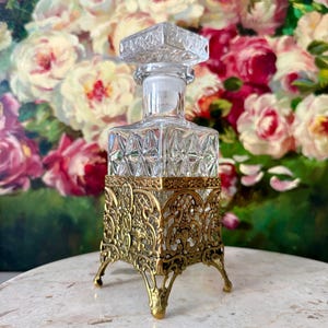 May include: A clear glass decanter with a square body featuring a diamond-cut pattern and a matching square stopper. The base of the decanter is encased in an ornate, gold-toned metal filigree stand with four decorative legs and intricate scrollwork.