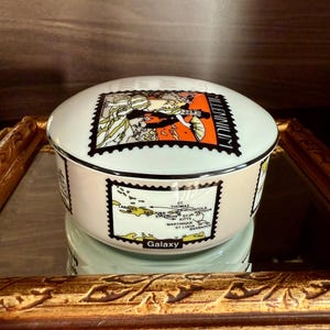 Peut inclure: Une boîte à bijoux ronde en céramique bleu clair avec un couvercle. La boîte est décorée de graphiques de style timbre-poste représentant une femme et une carte. Le mot "Millennium" est imprimé sur le couvercle. Le mot "Galaxy" est imprimé sur le côté.