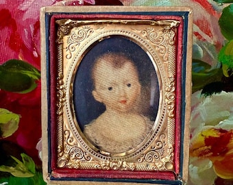 May include: A small, antique portrait of a baby in an oval frame. The frame is gold-colored with ornate detailing. The portrait is set against a dark blue background, with a red and blue border. The baby has fair skin and is wearing a light-colored garment.