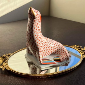 May include: A red and white porcelain figurine of a seal sitting on a gold-colored oval tray with a mirror surface. The seal is looking up and has a gold-colored detail on its head.