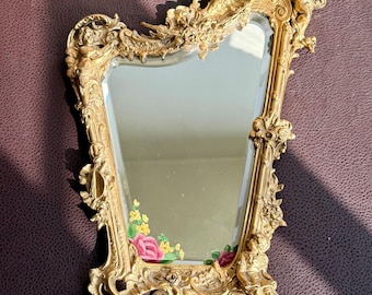 May include: Ornate gold-colored mirror with a decorative frame featuring cherubs, floral accents, and intricate detailing. The mirror's surface reflects a soft, neutral hue, and the bottom corners display painted pink roses and yellow flowers.