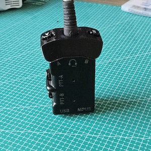 May include: A black electronic device with a USB port and the text "PTT-A" and "PTT-B". The device has a cable attached and is on a green grid surface. The device also has the text "MPUS".