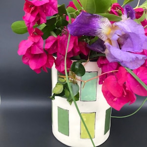 May include: A white ceramic vase with a geometric design of green rectangles holds a vibrant arrangement of pink bougainvillea, purple irises, and green foliage. The vase is cylindrical and sits on a dark surface.