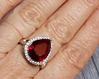 May include: A silver-toned ring featuring a large, pear-shaped, deep red gemstone as the centerpiece. The central stone is surrounded by a halo of small, clear, sparkling stones set into the metal band, creating a bright, contrasting border.