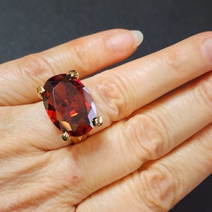 May include: A gold ring with a large, oval-cut, deep red gemstone. The gemstone is held in place by four prongs. The ring is shown on a hand against a dark background, highlighting the jewellery's design and colour.