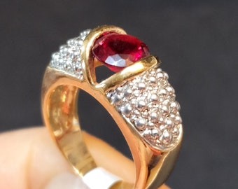 May include: A gold ring featuring a central oval-cut deep red gemstone. The band is constructed with a smooth gold finish on the lower portion and a textured, granular silver-toned surface on the upper shoulders surrounding the central stone.