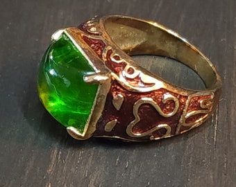 May include: A gold-toned ring featuring a large, square-cut, translucent green stone set in a prong setting. The band is decorated with deep red enamel inlay and raised gold-colored metal patterns that wrap around the sides of the ring.