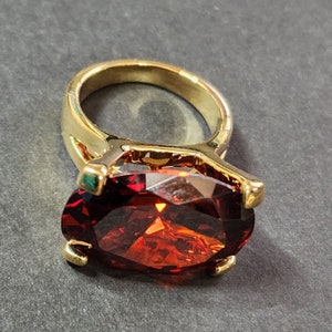May include: A gold-toned ring featuring a large, oval-cut, deep red gemstone. The ring's band is a simple, polished gold, and the gemstone is held in place by four gold prongs. The gemstone has multiple facets that reflect light.