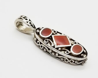 May include: Silver pendant with an oval shape, featuring a central square and two round, reddish-orange stones. The pendant has an ornate, scrollwork design around the stones and a loop for a chain.