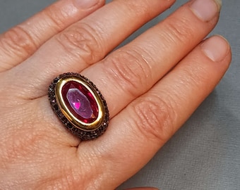 May include: An oval-shaped ring featuring a large, faceted, deep pink center stone set within a smooth gold-toned bezel. The surrounding band is wide and textured with a dark, metallic finish, creating a contrast against the bright center stone.