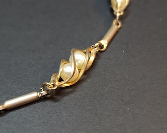 May include: Close-up of a gold-toned necklace with faux pearls. The necklace features a delicate design with two pearls encased in a swirling gold setting. The background is a dark, solid color.