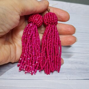 May include: A pair of vibrant hot pink tassel earrings constructed from numerous small glass beads. Each earring features a rounded, beaded top section that transitions into a long, flowing fringe of beaded strands hanging from a gold-tone hook.