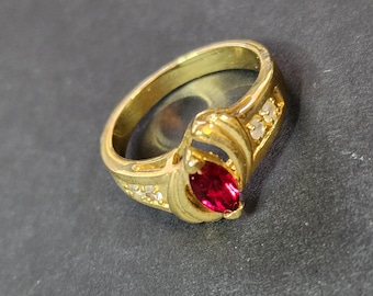 May include: A gold-toned ring featuring a central marquise-cut red gemstone. The band is accented with small, clear, round-cut stones embedded along the sides, creating a symmetrical design that highlights the vibrant red center stone.