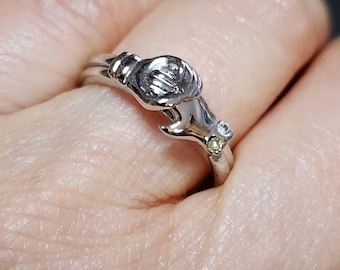 May include: A silver-toned ring featuring a detailed design of a hand holding a rose. The ring has a delicate, floral-inspired aesthetic, with small, clear and yellow gemstones adding sparkle.