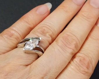 May include: A silver-toned ring featuring a single marquise-cut clear stone set within a bypass-style band. The metal band curves around the central gemstone, creating an open, flowing design that wraps across the finger with a polished finish.