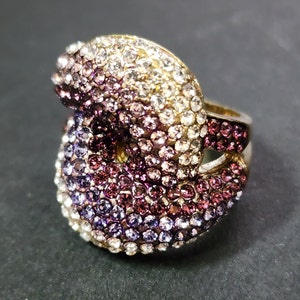 May include: A gold-toned ring encrusted with numerous small, sparkling crystals. The crystals transition in color from white to shades of purple and pink. The ring has a unique, intertwined design.