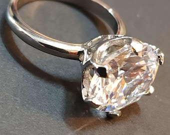 May include: A silver-toned ring with a large, clear, faceted gemstone. The ring's band is smooth and polished, and the gemstone is held in place by prongs. The ring is set against a dark background.