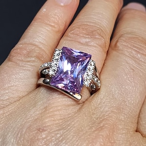 May include: A silver ring featuring a large, rectangular, purple gemstone surrounded by smaller clear gemstones.