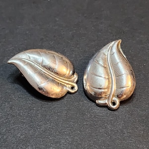 May include: A pair of silver-toned leaf-shaped earrings. Each earring has a detailed vein pattern and a small loop at the base. The earrings are set against a dark background.
