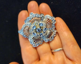 May include: A silver-toned flower brooch with a detailed openwork design. The brooch is adorned with numerous light blue rhinestones, with larger blue stones at the center. The floral design is set against a dark background.