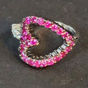 May include: A silver ring with a heart-shaped design. The heart is outlined with small, bright pink gemstones. The ring has a detailed band with additional small gemstones. The ring is set against a dark background.