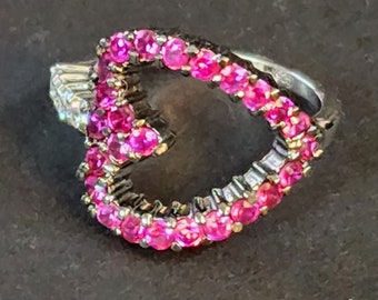 May include: A silver ring with a heart-shaped design. The heart is outlined with small, bright pink gemstones. The ring has a detailed band with additional small gemstones. The ring is set against a dark background.