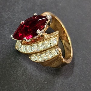 May include: A gold-toned ring featuring a large, marquise-cut deep red stone set in silver-colored prongs. The band is accented with two parallel rows of small, clear, round-cut stones that follow the curve of the ring's wide, metallic shank.