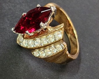 May include: A gold-toned ring featuring a large, marquise-cut deep red stone set in silver-colored prongs. The band is accented with two parallel rows of small, clear, round-cut stones that follow the curve of the ring's wide, metallic shank.
