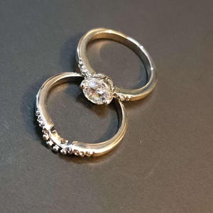 May include: Two silver rings with a single clear stone on the top ring. The bottom ring has a decorative edge.