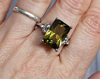May include: A silver ring with a large, rectangular, olive-green gemstone as the focal point. The ring is accented with smaller, clear gemstones clustered around the main stone. Another silver ring is visible on the adjacent finger.