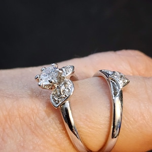 May include: A silver engagement ring set with a large, round, clear center stone and smaller stones set in a floral design. The ring is worn on a finger.