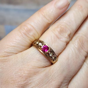 May include: A gold ring featuring a vibrant pink gemstone at its center, flanked by smaller, clear stones. The band has an intricate, openwork design. The ring is worn on a finger, showcasing its elegant details and craftsmanship.