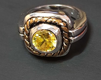 May include: A silver and gold ring with a yellow gemstone set in a square bezel. The ring has a braided design around the bezel.
