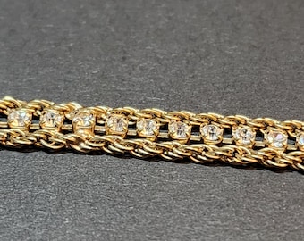 May include: Close-up of a gold-toned bracelet with a braided design. The bracelet features a row of clear, sparkling gemstones set between the braided sections. The background is a dark gray.
