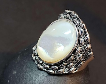 May include: A ring featuring a large, oval-shaped, iridescent white mother-of-pearl cabochon set in a dark, oxidized silver-toned metal band. The band has a highly textured, bumpy, and irregular surface that surrounds the central stone.