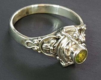 May include: A silver ring with ornate detailing and a round, light green gemstone. The ring's design features floral and scrollwork patterns, with the gemstone set in a bezel setting. The ring is shown against a dark background.