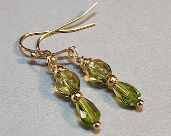May include: A pair of dangle earrings featuring faceted, light green beads and gold-colored metal accents. The earrings have a hook closure and a teardrop-shaped bead at the bottom. The earrings are displayed on a light gray surface.