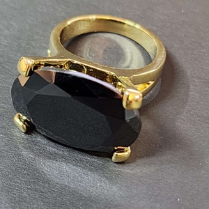 May include: A gold-colored ring featuring a large, faceted black gemstone. The gemstone is oval-shaped and securely held in place by four gold prongs. The ring band is a simple, polished gold.