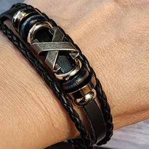 May include: Black leather bracelet with a braided design and silver-tone accents. The bracelet features a silver-tone X-shaped centerpiece and gold-tone metal rings. The bracelet is multi-layered and appears to be worn on a wrist.