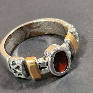 May include: Silver ring with gold accents and a deep red, oval-shaped gemstone. The ring features intricate detailing on the band and gold rectangular inlays. The gemstone is set in a silver bezel, creating a striking contrast.