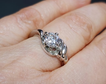May include: A silver ring with a large, round, clear gemstone. The band has a swirling design that wraps around the stone. The ring is on a finger, with the skin visible in the background.