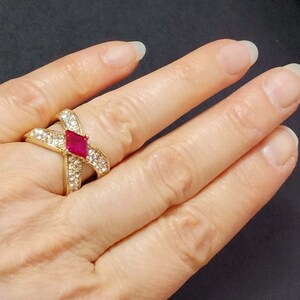 May include: A gold-toned crossover ring featuring a central marquise-cut vibrant red stone flanked by two bands encrusted with numerous small clear white stones. The design forms an X-shape across the finger, showcasing a polished metal finish.